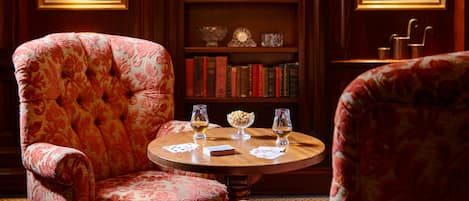 Una habitación con una mesa redonda de madera, dos sillones, una estantería con libros y objetos decorativos, y cuadros en las paredes.