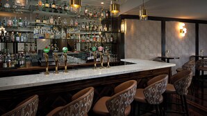 Zona de bar con varias estanterías llenas de botellas de licor, mostrador de bar con grifos y varios taburetes con diseños.