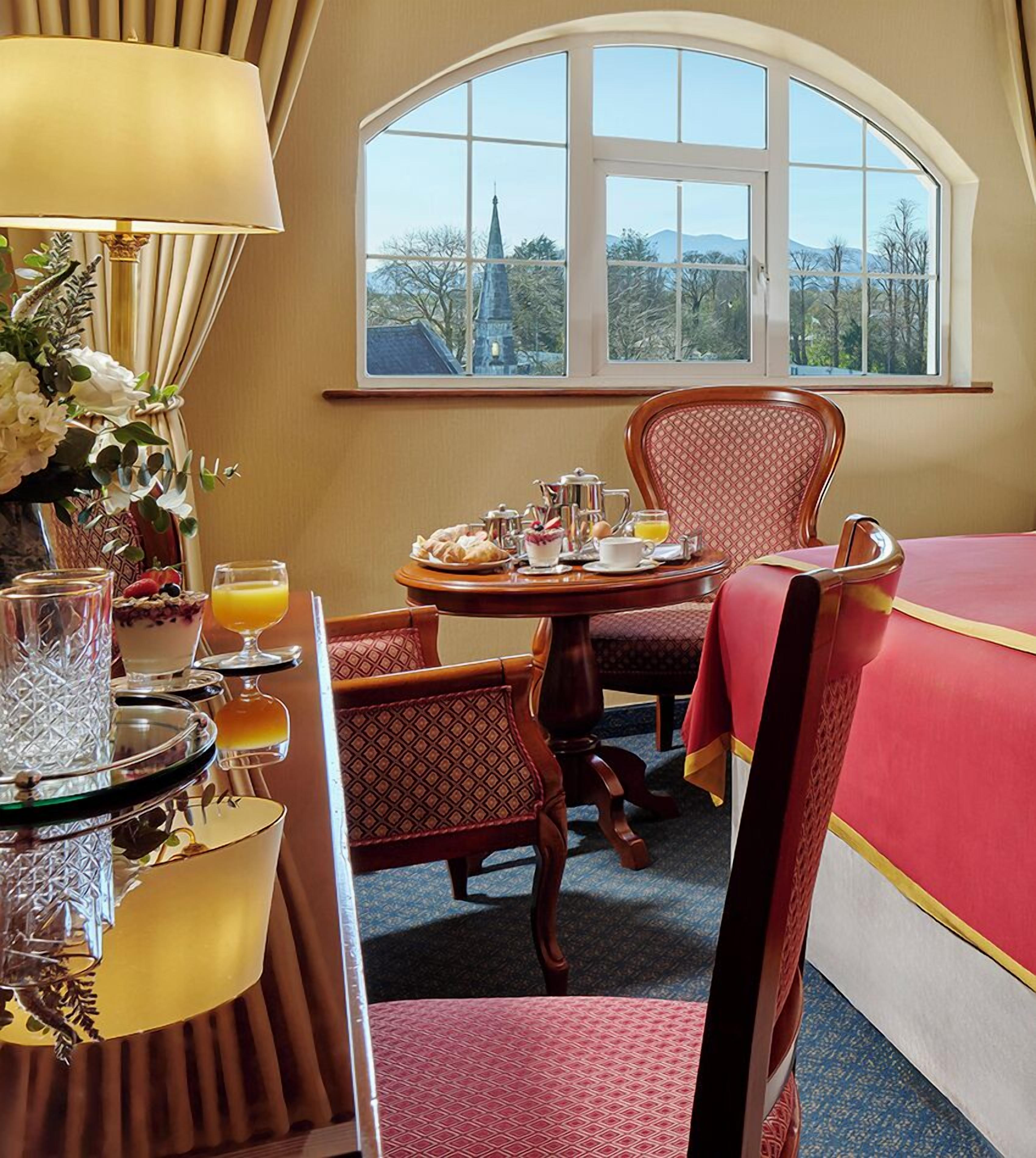 Habitación de hotel con una mesa de comedor lista para el desayuno, una cama con ropa de cama roja y un ventanal con vistas a una iglesia y montañas.