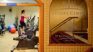 Zona de gimnasio con equipo de ejercicio y una persona usando una cinta de correr. Hay un cartel del gimnasio y una ventana con el texto "KTH Leisure Centre O'Donoghue Hotels".