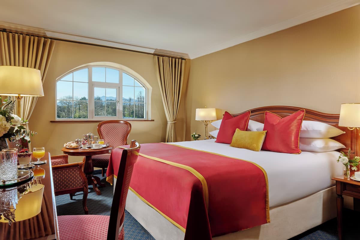 Ein Hotelzimmer mit einem großen Bett, einem Esstisch mit Stühlen, einem kleinen Tisch mit Lampe und einem Fenster mit Vorhängen.