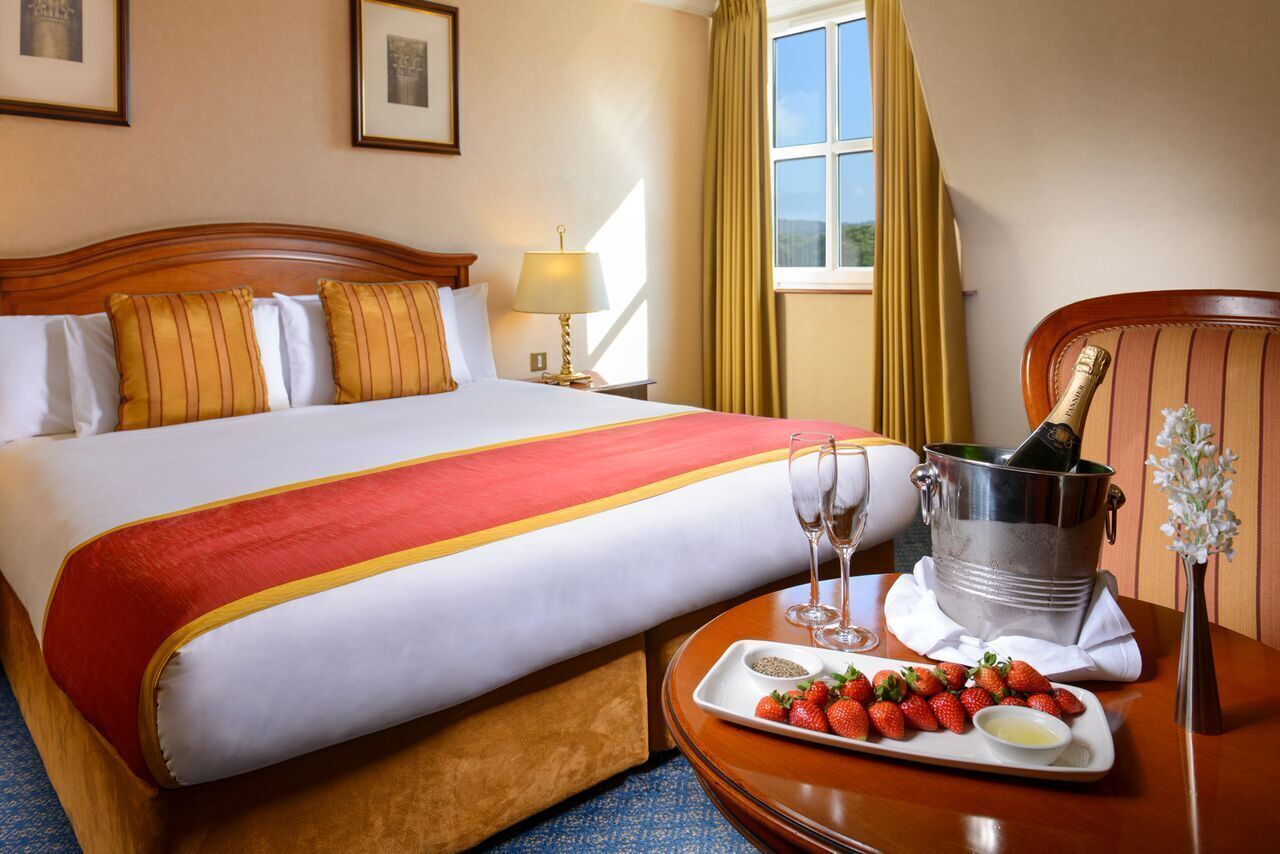 Habitación de hotel con cama, una bandeja de fresas y champán, y una mesita de noche con lámpara.