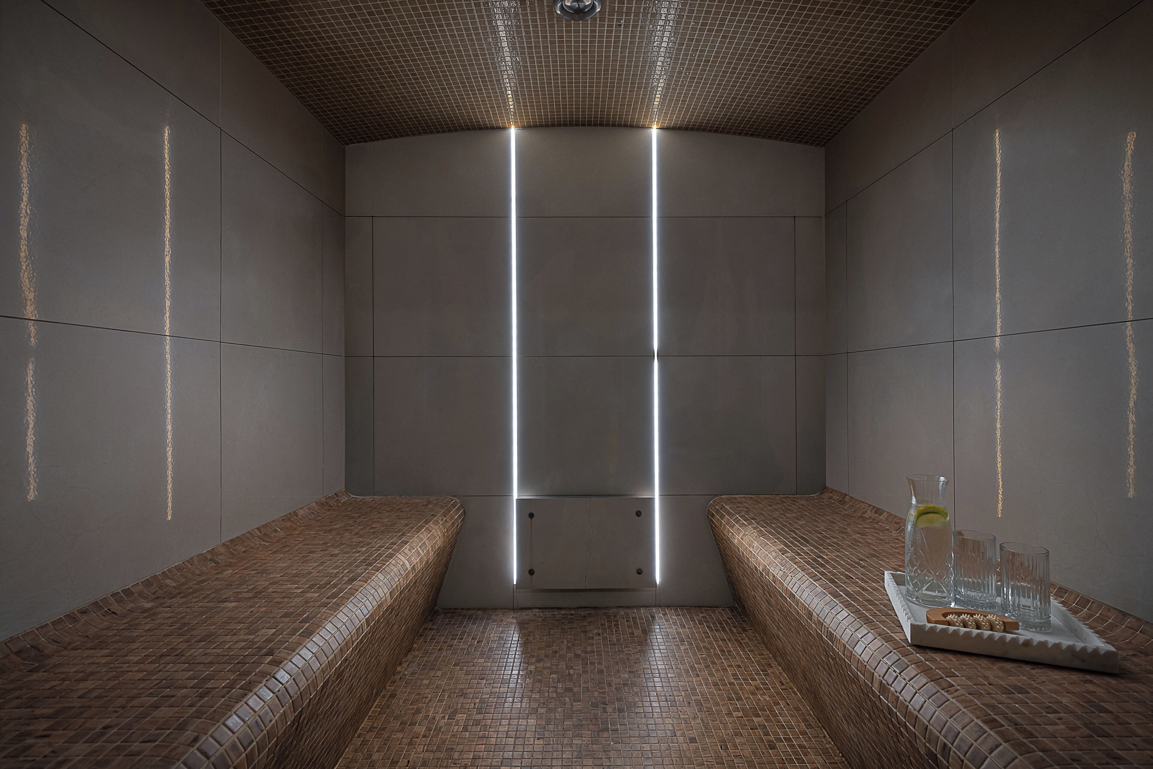 Un cuarto de spa con suelo de baldosas, bancos, una bandeja con vasos y una botella, y un techo con diseño de cuadrícula.