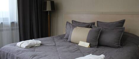 Una cama bien hecha con edredón gris, dos toallas blancas y varias almohadas.