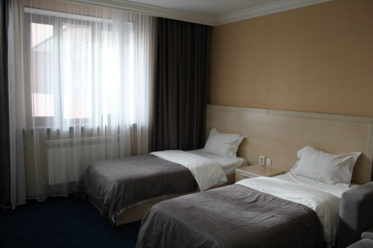 Habitación de hotel con dos camas individuales, una mesita de noche, una lámpara y un ventanal con cortinas traslúcidas.