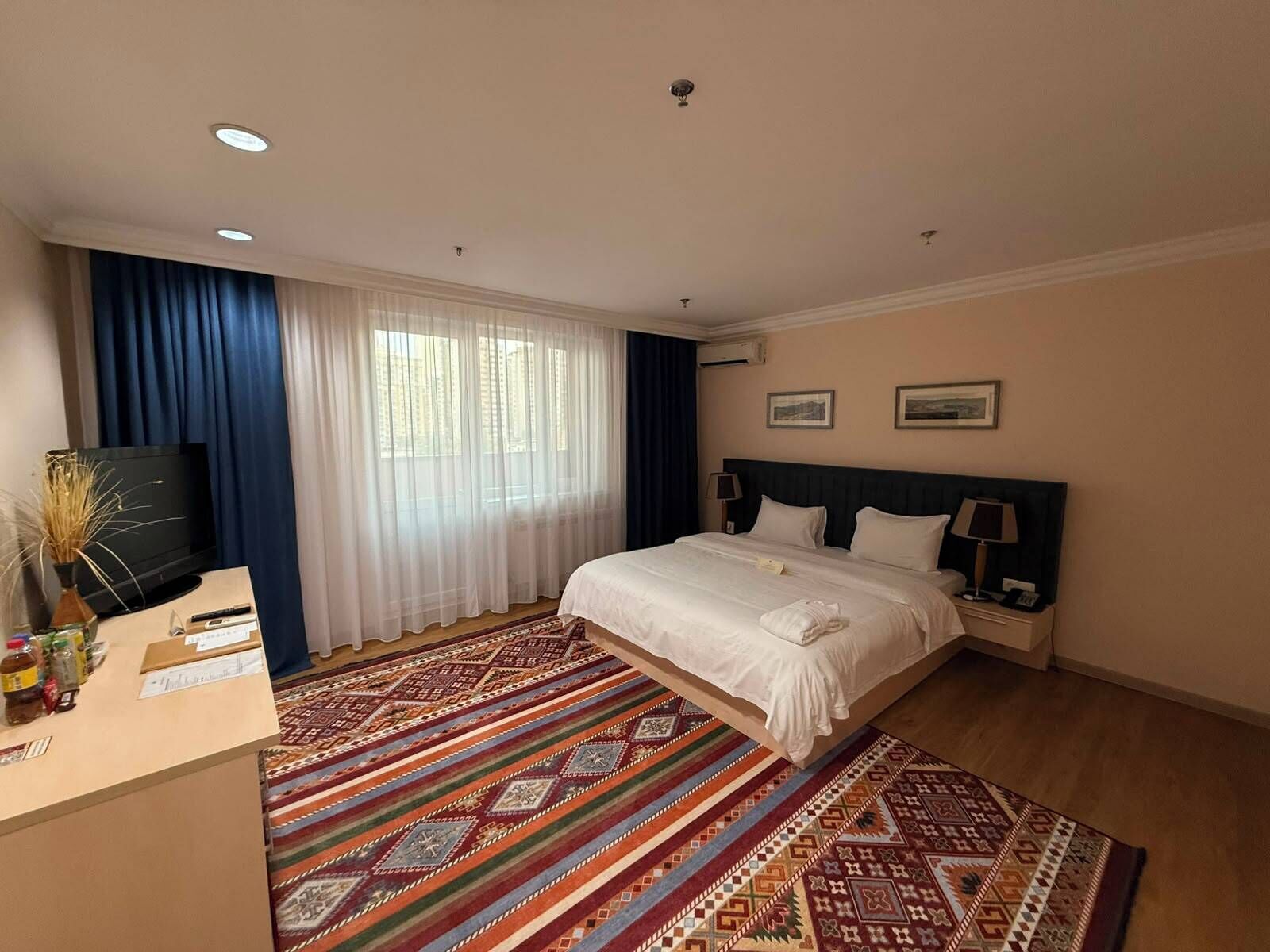 A hotel room with a bed, a desk with a computer, a television, and a patterned rug.