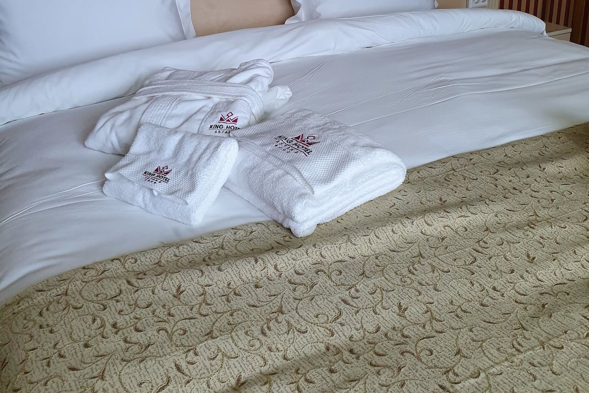Una cama bien hecha con sábanas blancas y dos almohadas, una alfombra estampada y un papel tapiz a rayas al fondo.