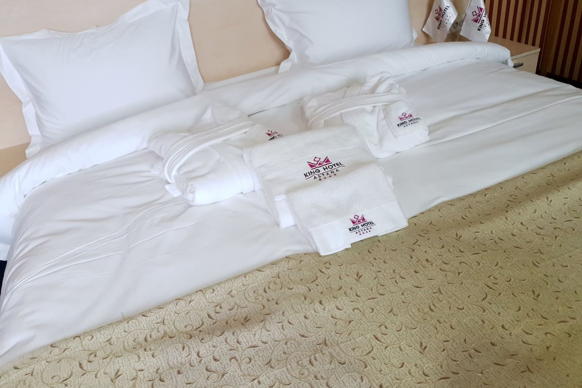 Una cama bien hecha con sábanas blancas, una colcha texturizada color beige y toallas con el logotipo del Hotel King.