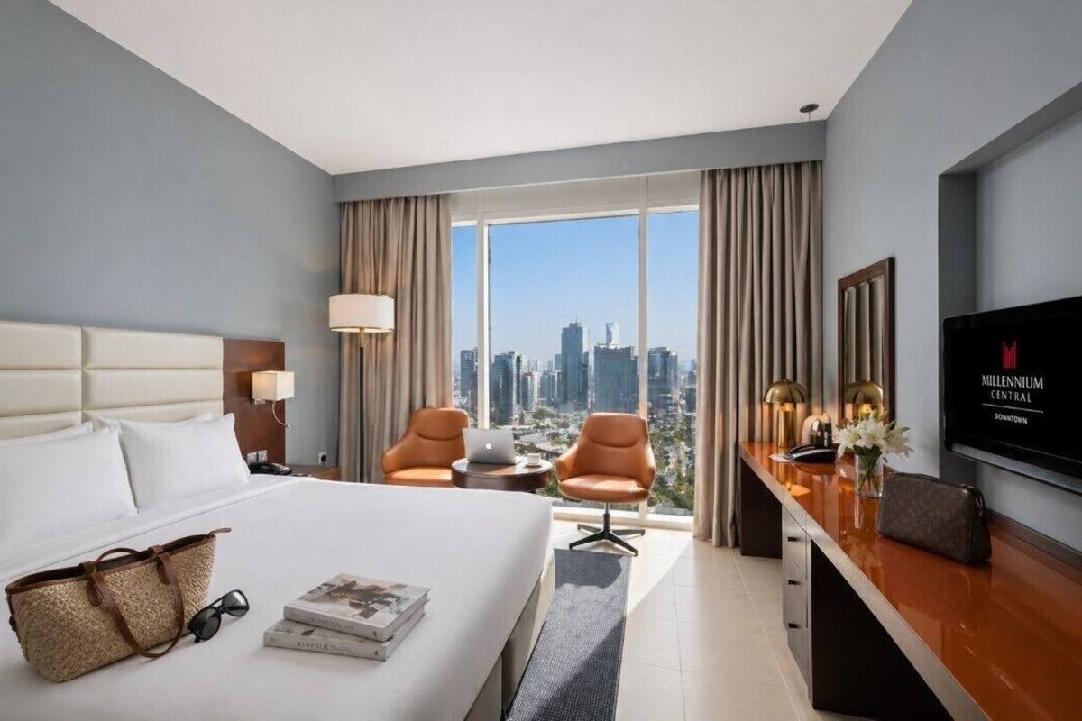 A hotel room with a bed, a desk, two chairs, a TV, and a view of the cityscape.
