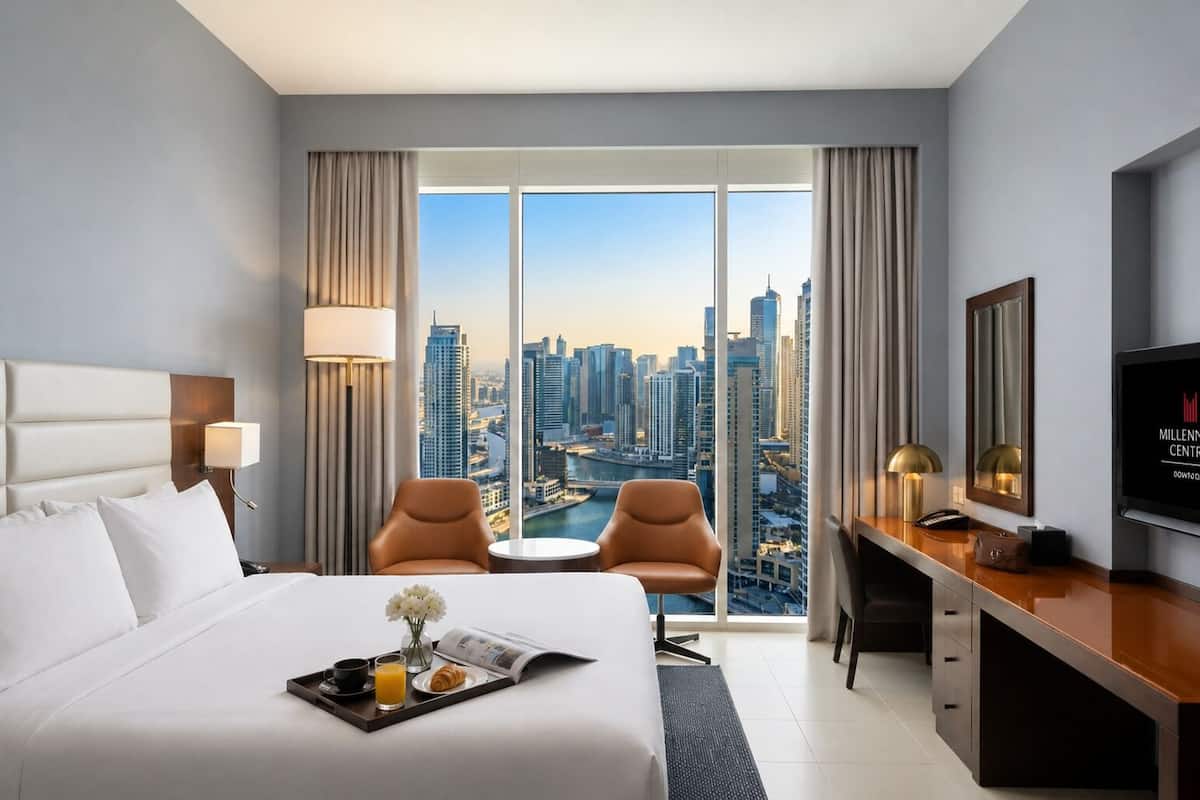 A hotel room with a bed, two chairs, a desk, a TV, and a large window with a city view.