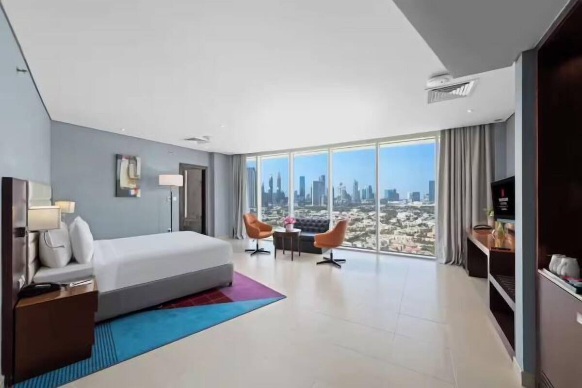 A modern hotel room with a large bed, a desk, two chairs, a TV, and a city view.