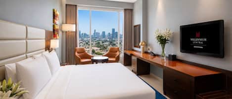 A hotel room with a large bed, a desk, two chairs, a TV, and a city view.