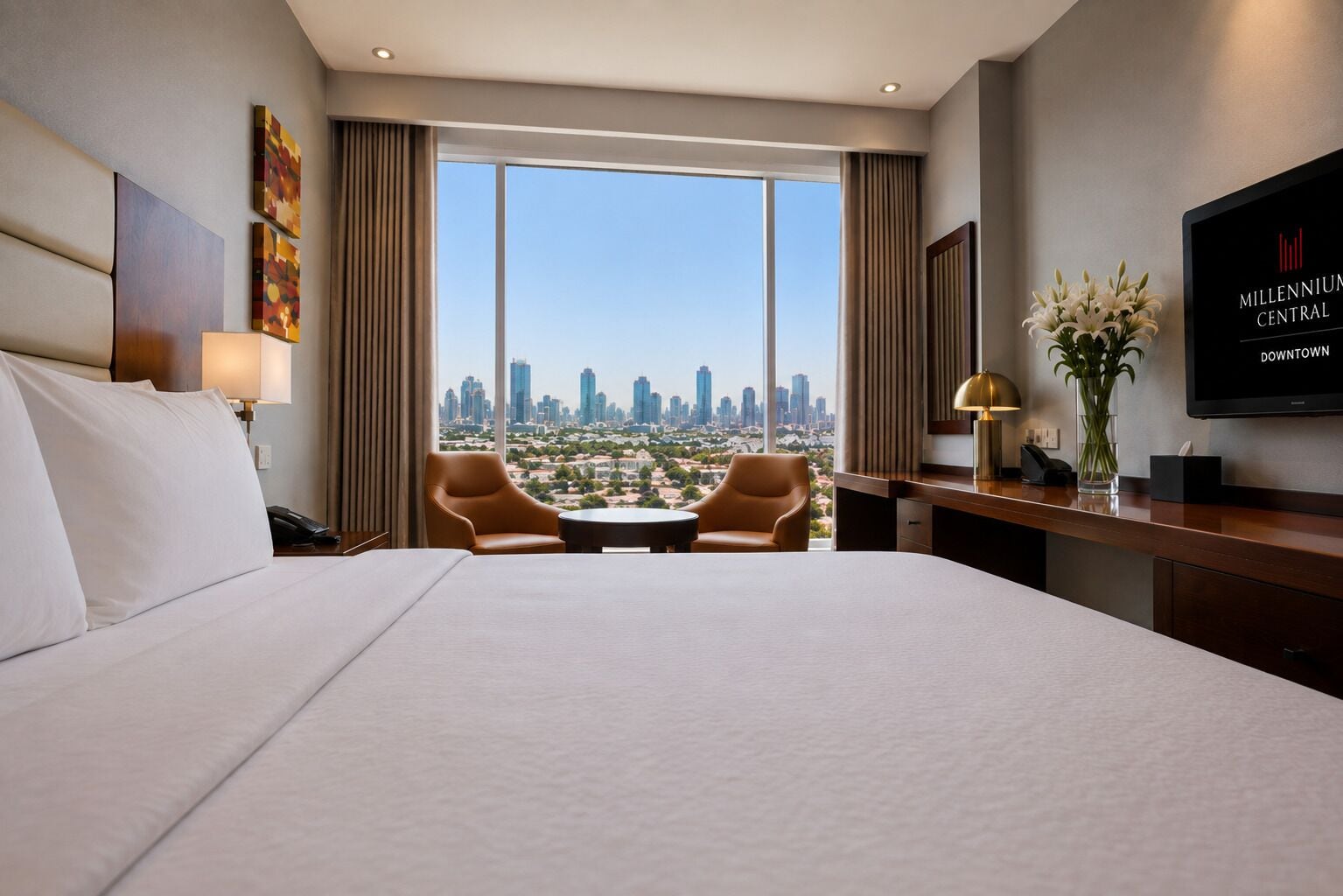 A hotel room with a large bed, two armchairs, a desk, a TV, and a view of the city skyline.