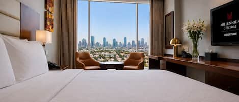 A hotel room with a large bed, two armchairs, a desk, a TV, and a view of the city skyline.