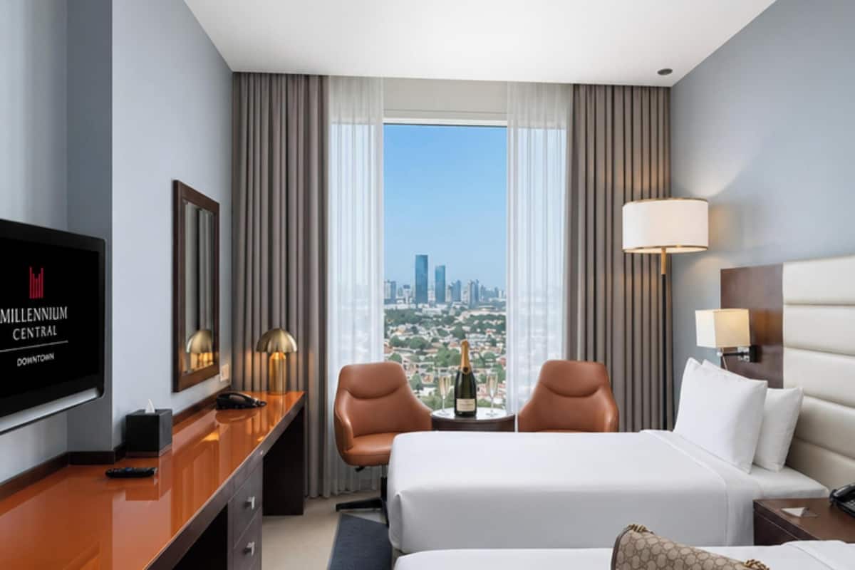 A hotel room with a bed, a desk with a TV, a chair, a table with a bottle, and a view of the city.