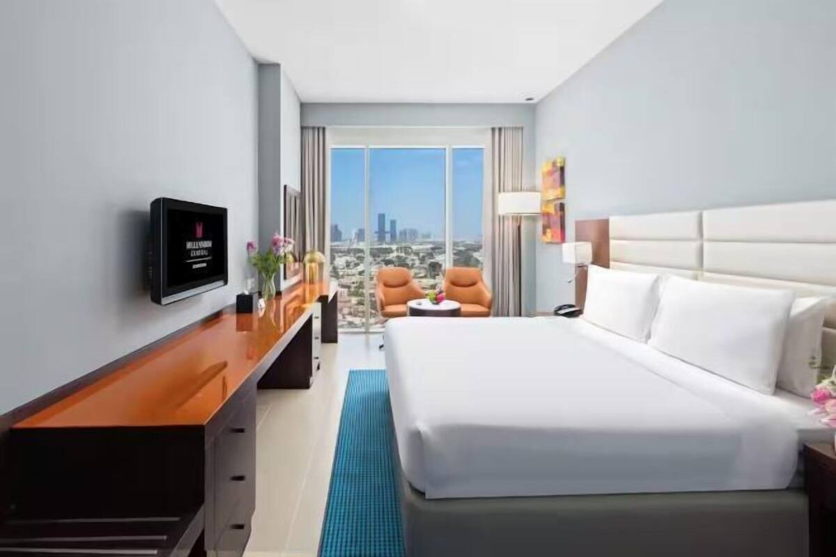 A hotel room with a bed, a desk with a TV, a chair, a small table, and a view of the city.