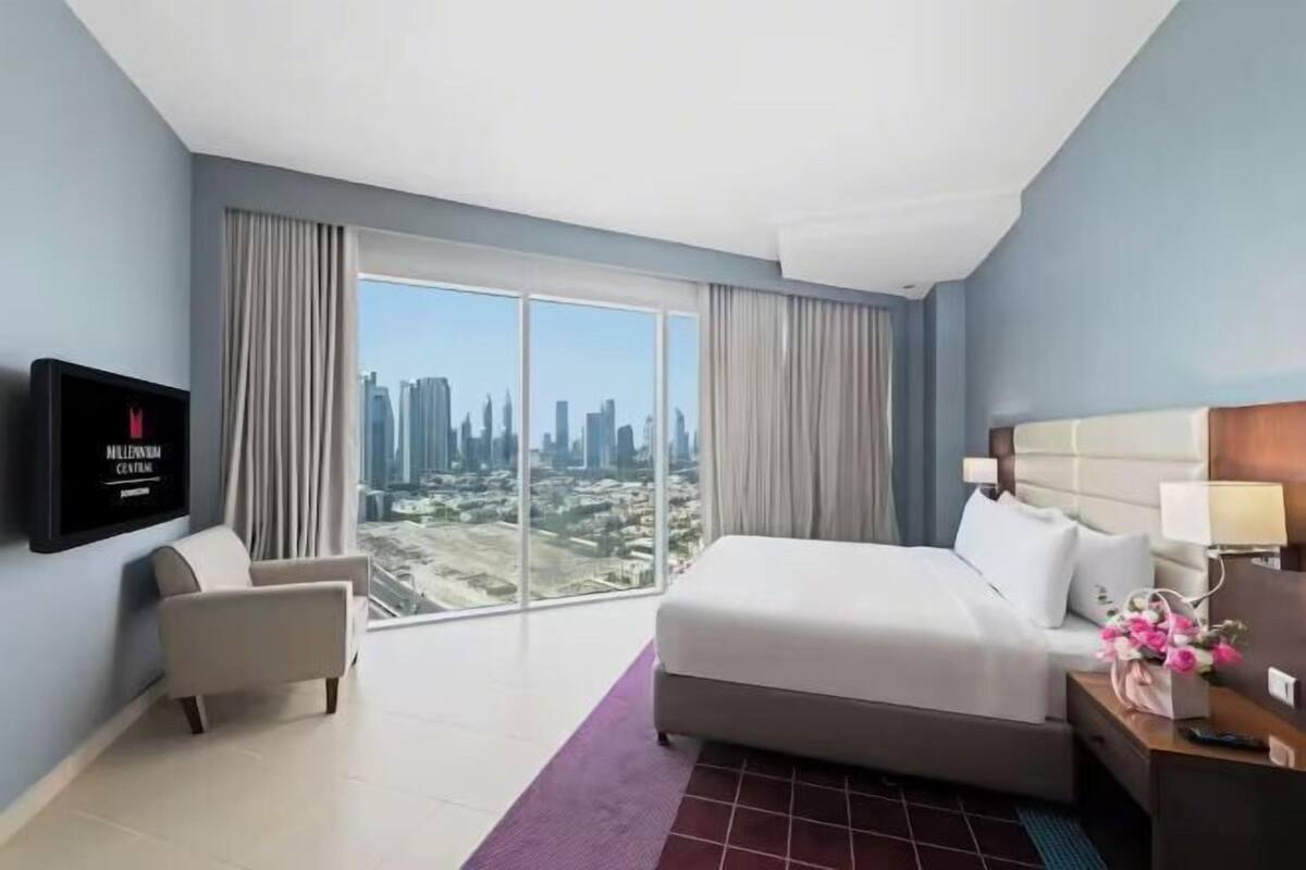 A modern hotel room with a large bed, a TV mounted on the wall, a chair, and a view of a cityscape through large windows.