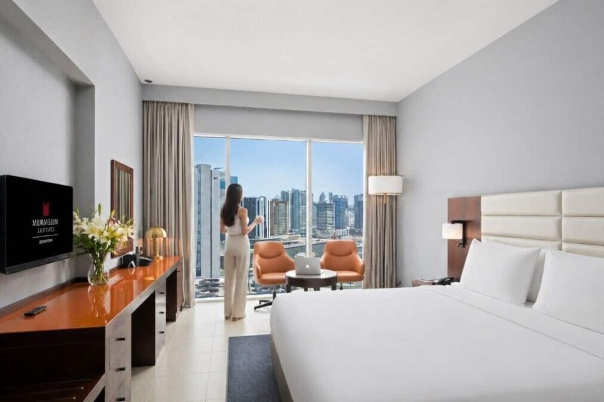 A hotel room with a bed, a desk with a laptop, two chairs, a TV, and a view of the cityscape.