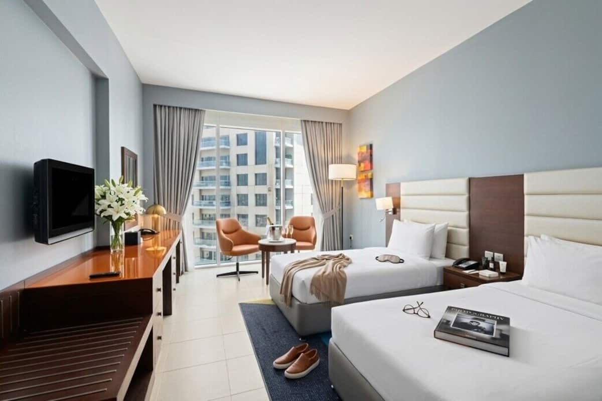 A hotel room with a bed, bedside table, TV, seating area with chairs, and a window with a view of buildings.