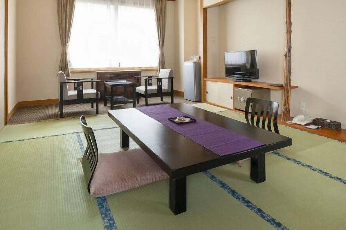 A room with a wooden ceiling, a dining table with chairs, a television, and a small kitchenette.
