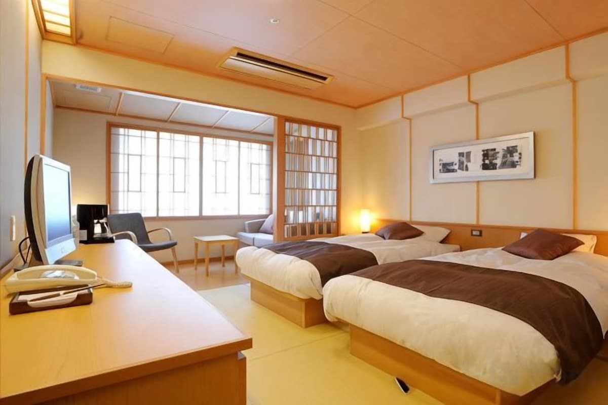Una habitación de hotel con escritorio, dos camas, una silla, una mesita y una estantería.