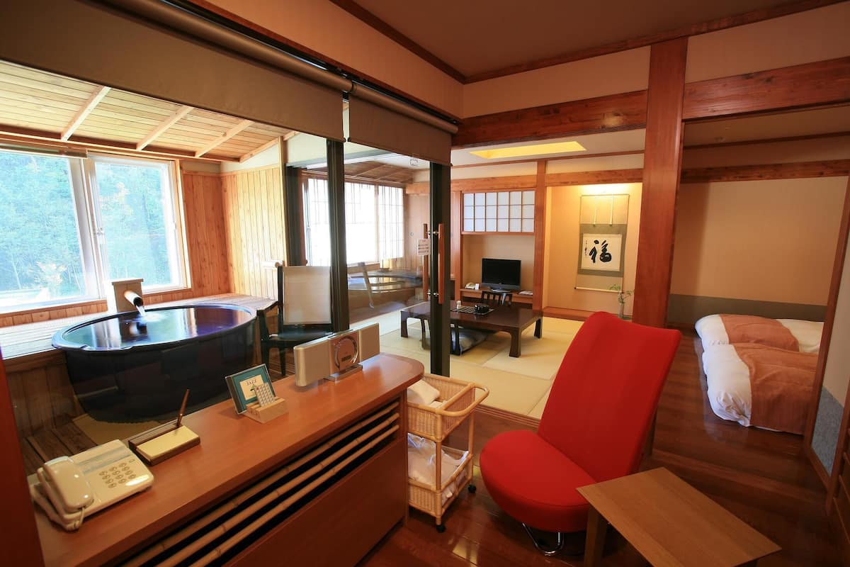 A traditional Japanese room with tatami flooring, a wooden desk with a telephone, a red chair, a bed with a futon, and a television.