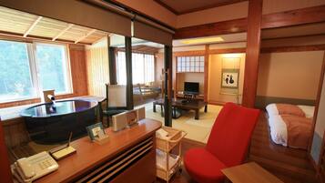 A traditional Japanese room with tatami flooring, a wooden desk with a telephone, a red chair, a bed with a futon, and a television.