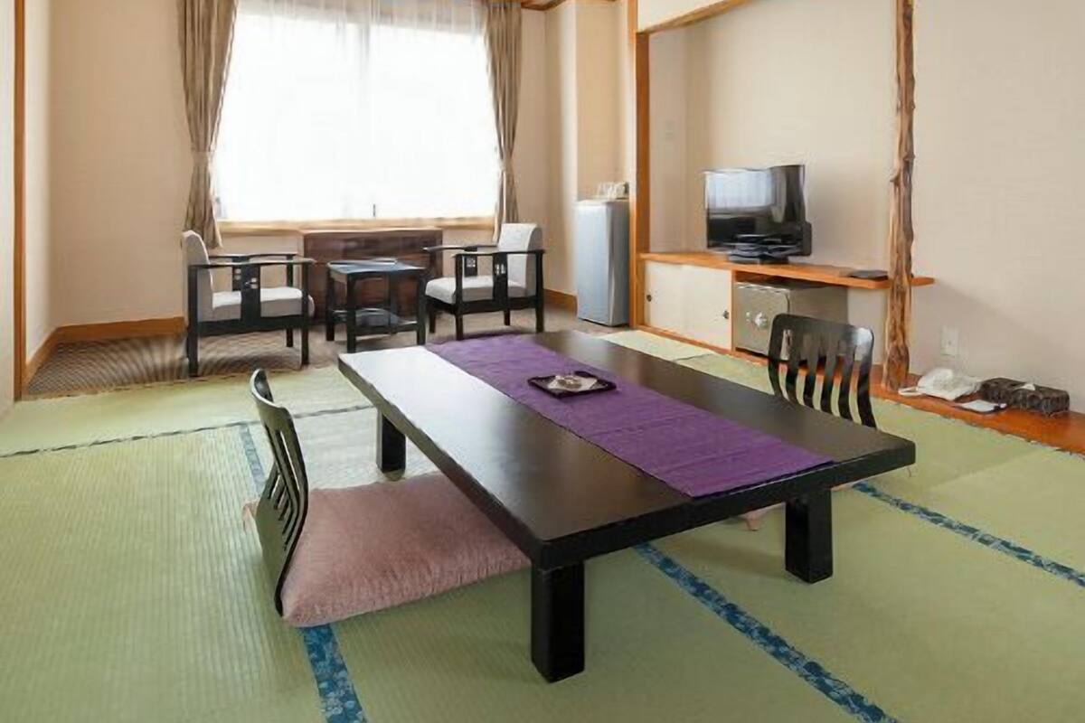 A room with a tatami mat floor, a low table with chairs, a television, and a window with curtains.
