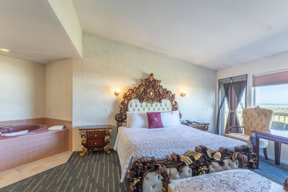 A hotel room with a large bed, a sitting area with a chair, a small table, a bathtub, and a view of the outdoors.