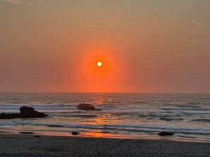 Una playa con el sol poniéndose sobre el océano, rocas en el agua y algunas aves en la arena.