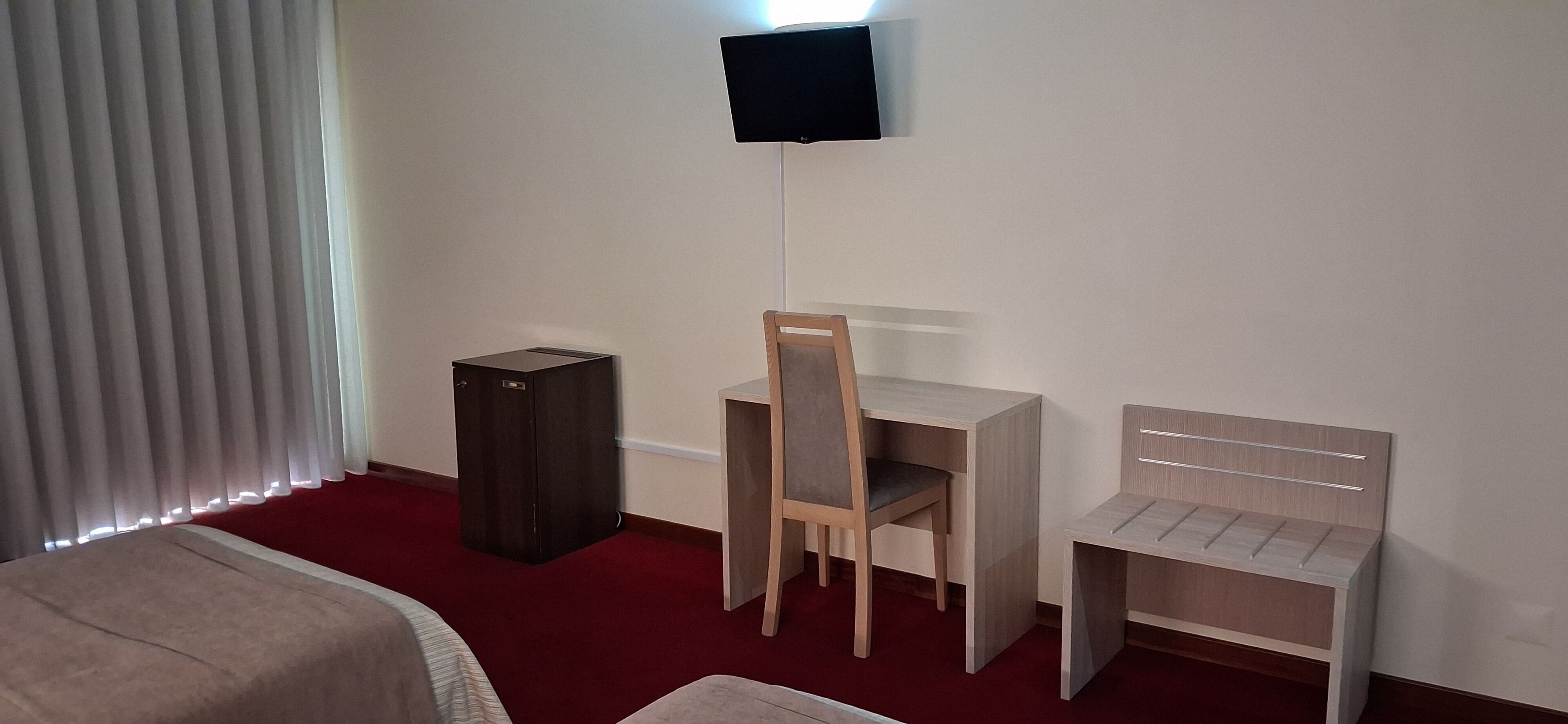 A hotel room with a bed, a desk, a chair, a small table, a TV mounted on the wall, and a tall floor lamp.