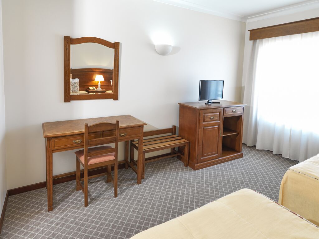 A hotel room with a wooden desk, a chair, a television, a wardrobe, and a bed.