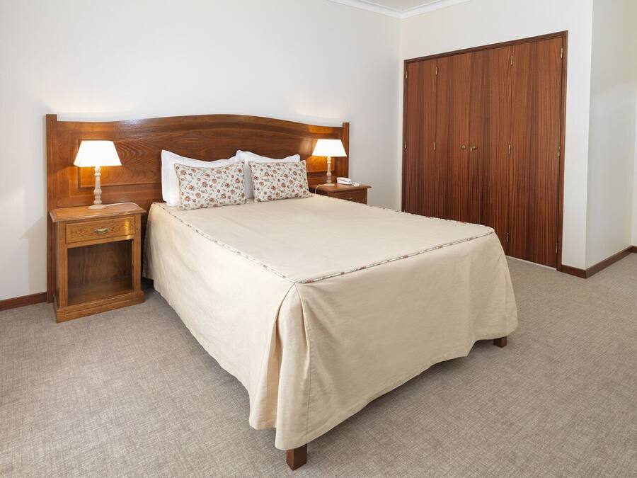 Un dormitorio con cabecera de madera, una cama con colcha color beige, dos mesitas de noche con lámparas y un armario de madera.