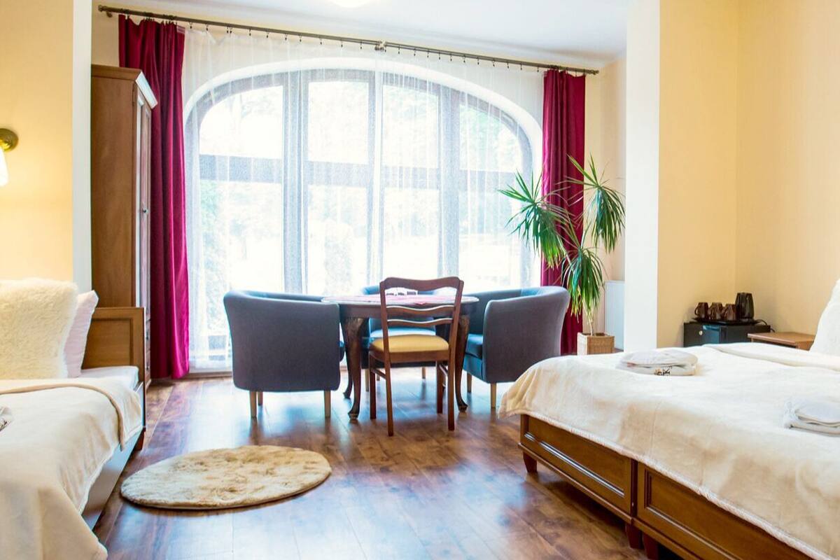 Een hotelkamer met twee bedden, een eettafel, stoelen, een kamerplant en een groot raam met gordijnen.