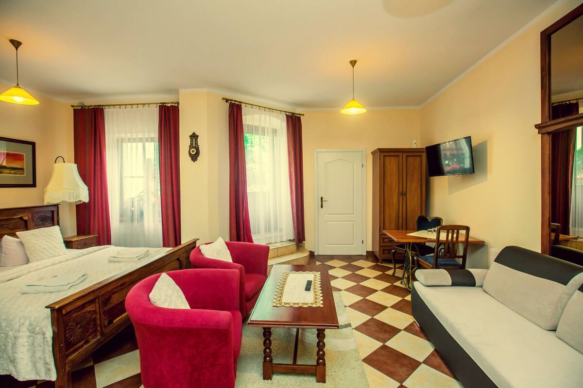 Een hotelkamer met een bed, een rode fauteuil, een bank, een houten tafel, een televisie en een geruit vloerkleed.