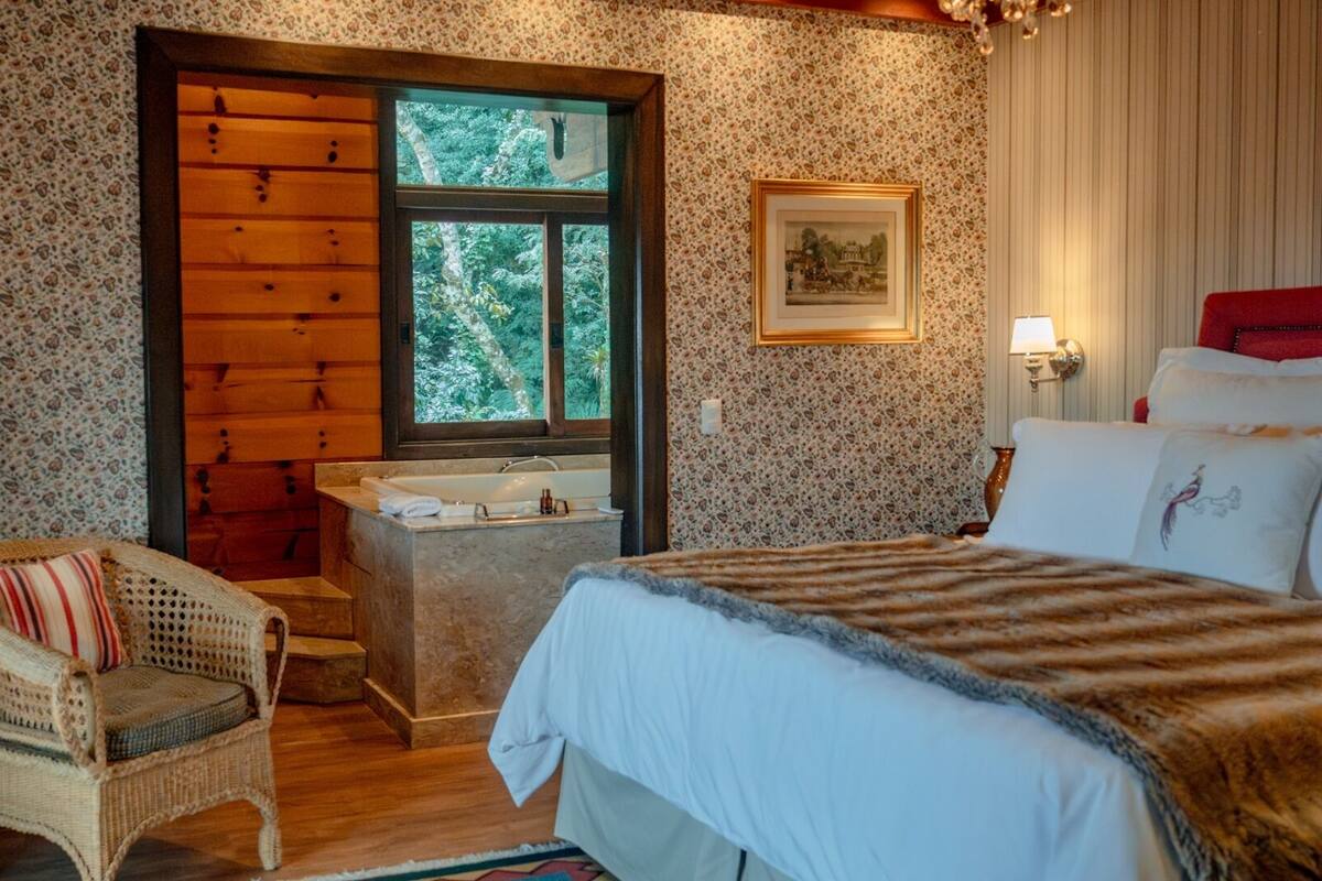 A cozy bedroom with a wooden door, a large window, a bed with a striped blanket, a wicker chair, and a small bathroom with a bathtub.