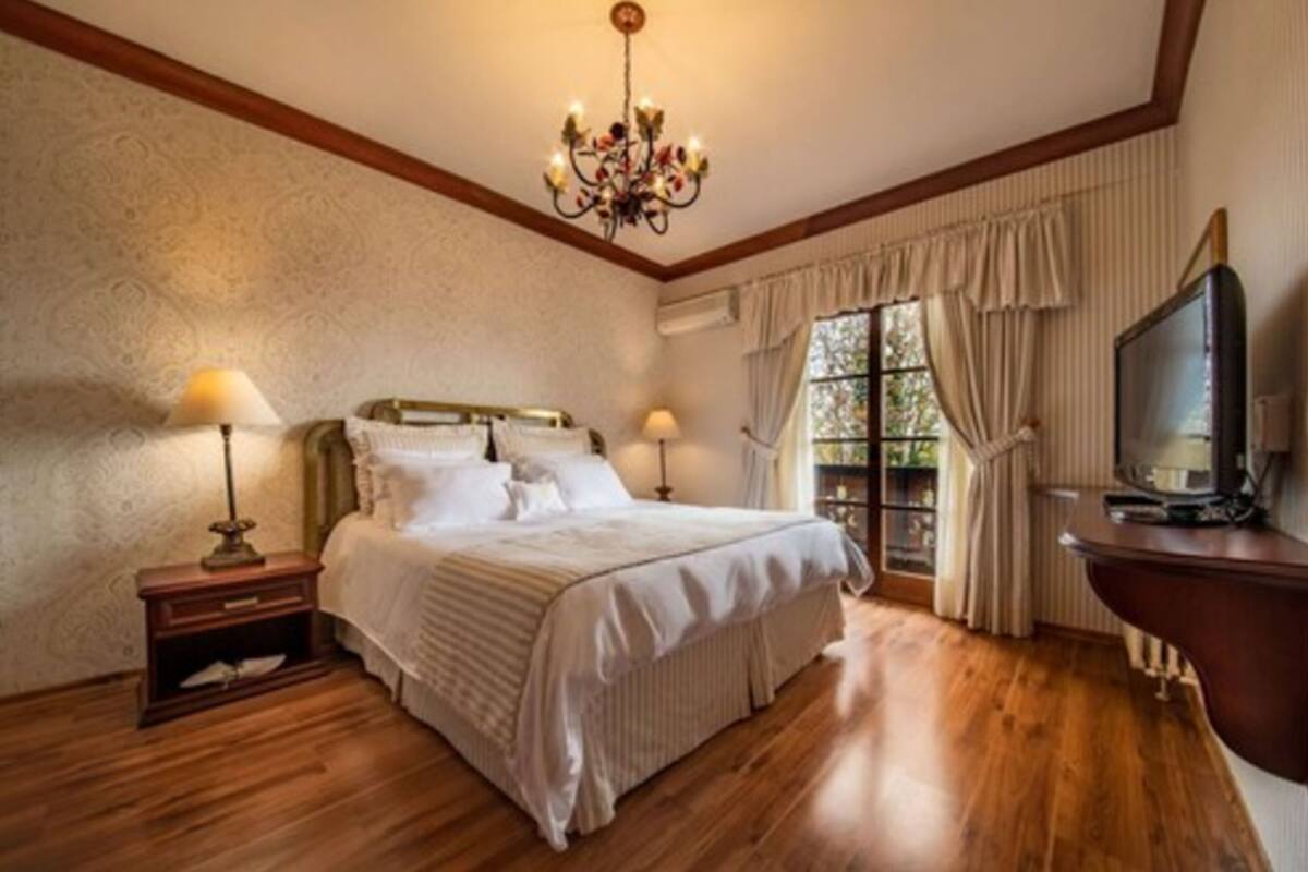 A bedroom with a bed, two bedside tables, a chandelier, a television, and a window with curtains.