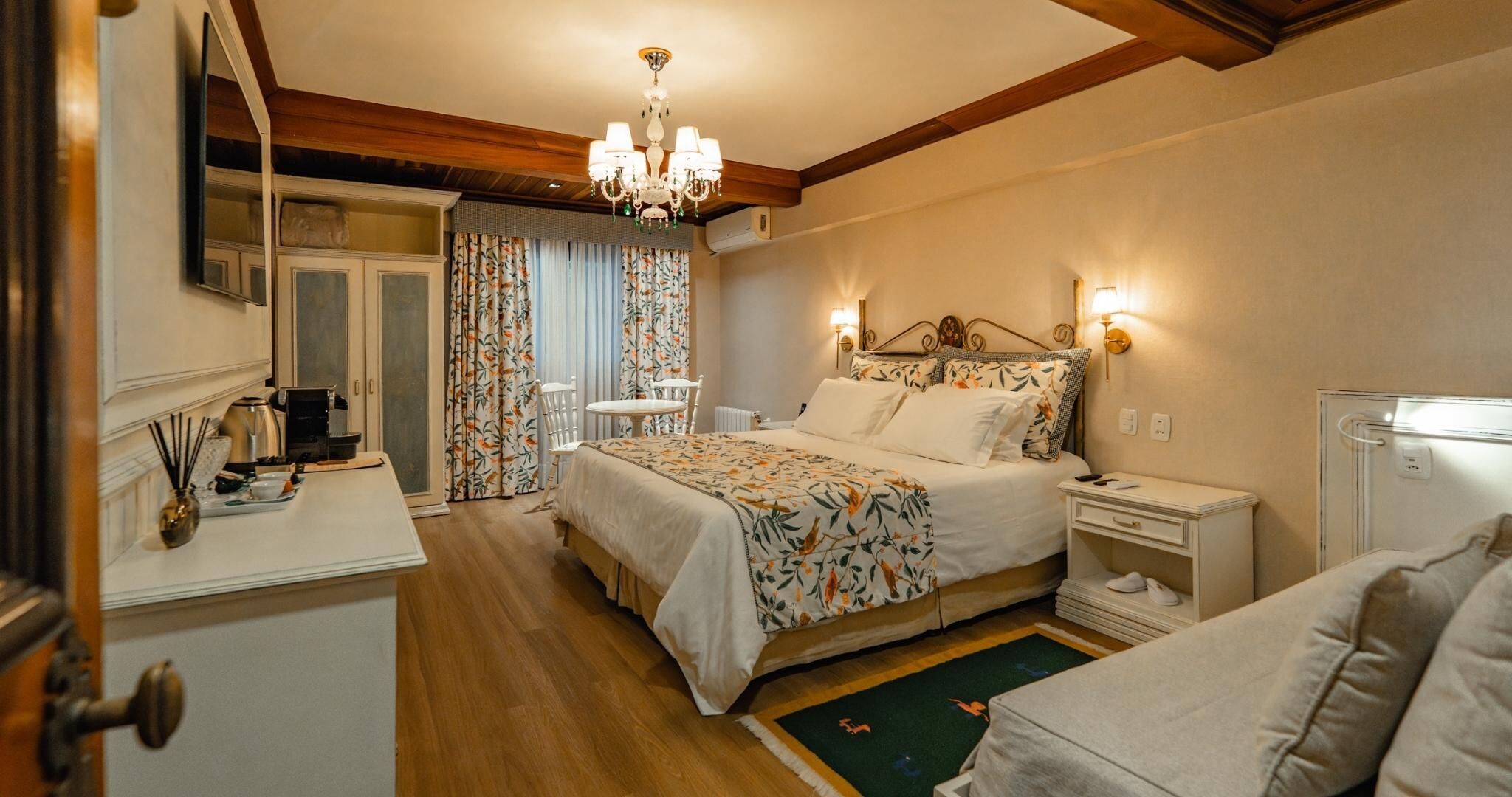A hotel room with a bed, bedside tables, a desk with a chair, a chandelier, and a mirror.