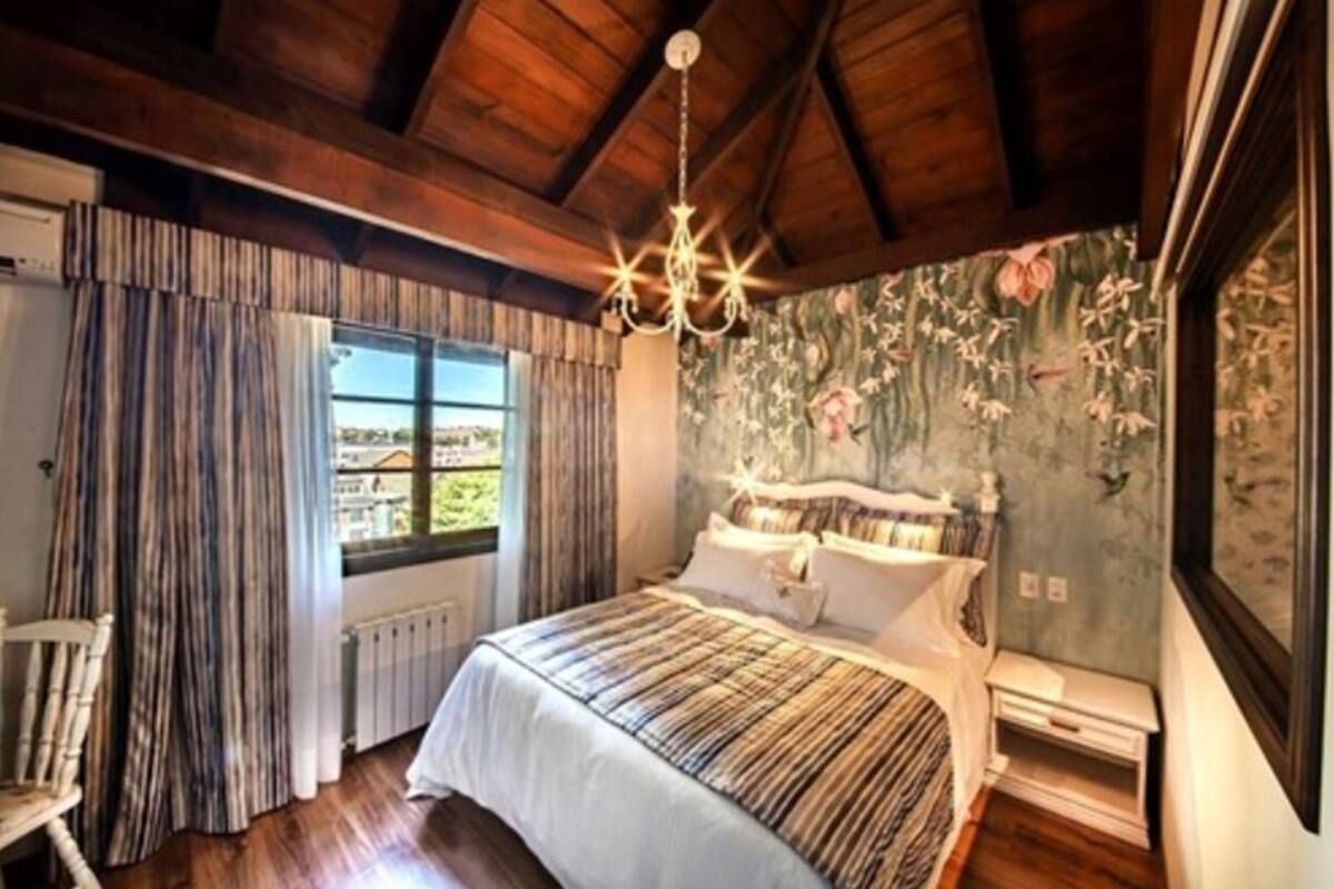 A bedroom with a large bed, a chandelier, a window with curtains, and floral wallpaper.