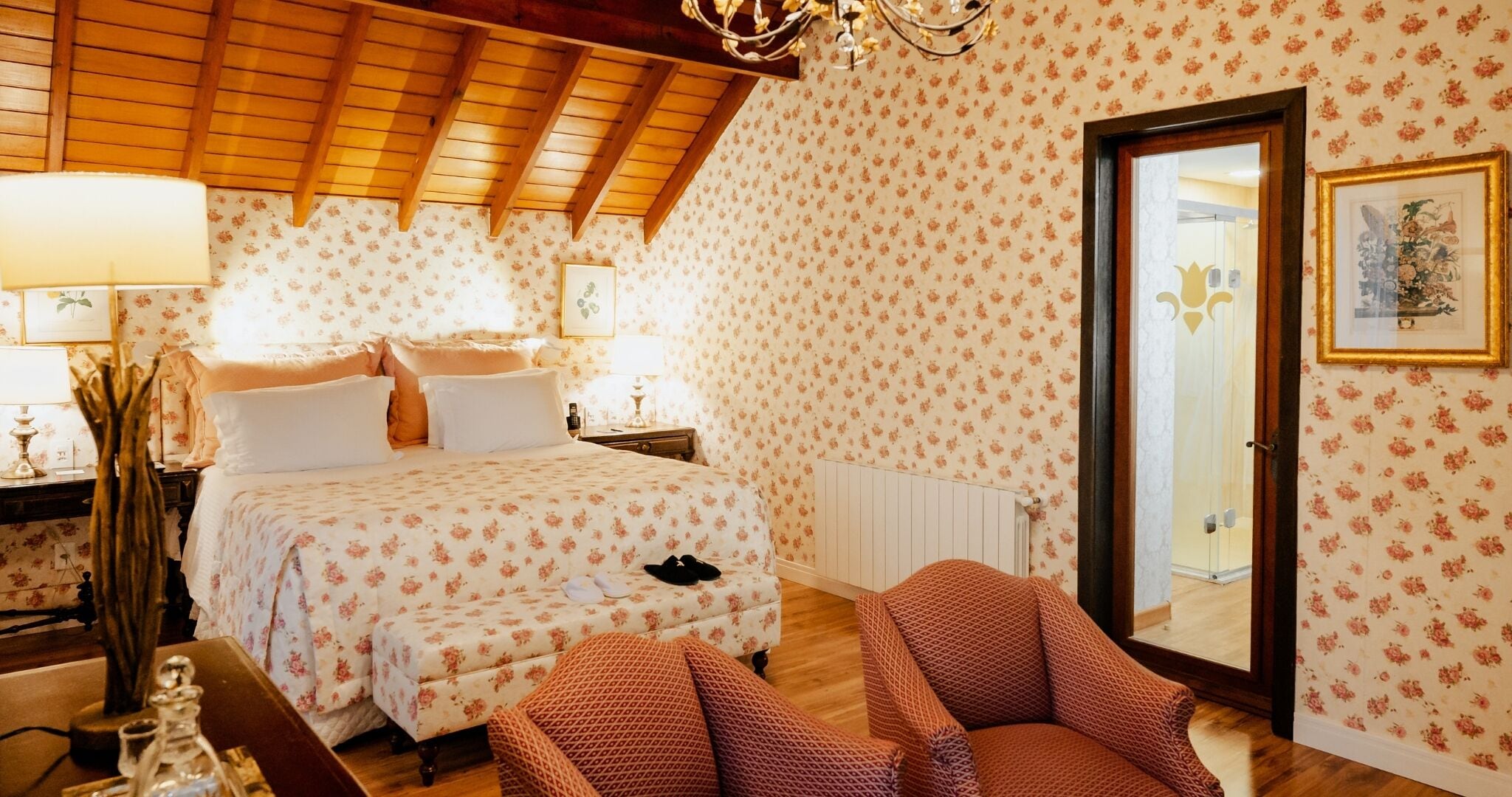 Un dormitorio con techo de madera, papel tapiz floral, cama con ropa de cama blanca y rosa, mesita de noche, lámpara y una silla.