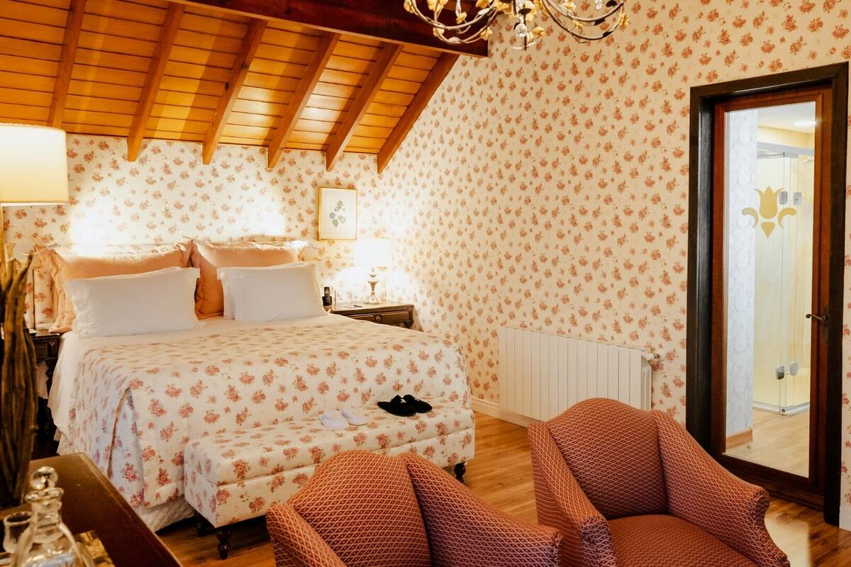 A bedroom with a wooden ceiling, floral wallpaper, a bed with white and pink bedding, a nightstand, a lamp, and a chair.
