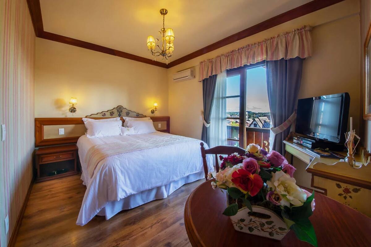 A hotel room with a bed, bedside lamps, a TV, a wooden desk, and a window with curtains.