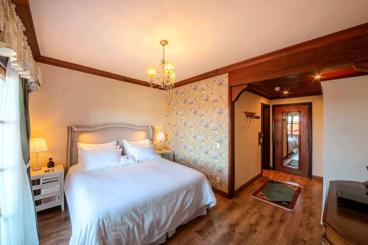 A bedroom with a large bed, a chandelier, floral wallpaper, wooden flooring, and a door leading to another room.