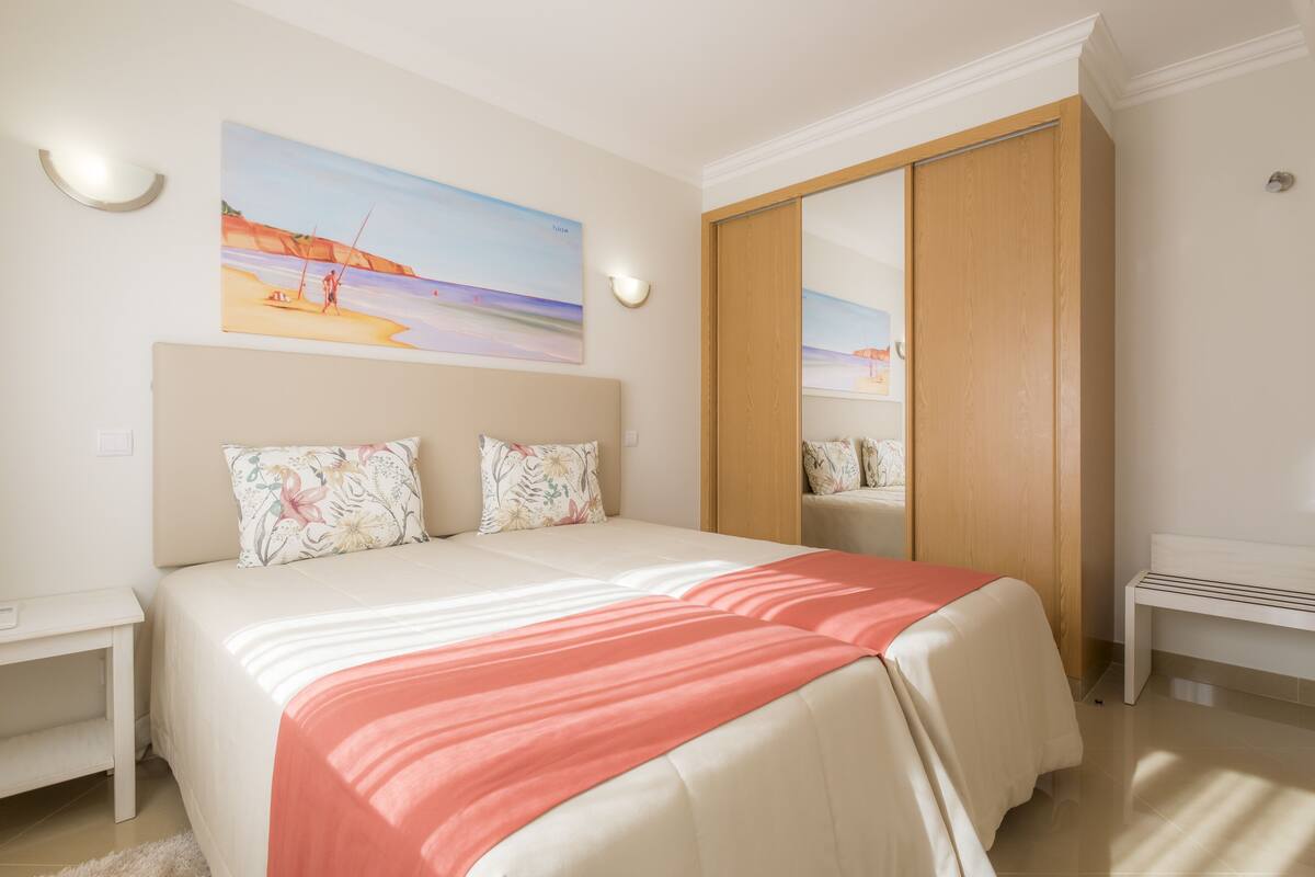 Een slaapkamer met een bed, een nachtkastje, een bankje, een kledingkast en een schilderij van een strandgezicht.