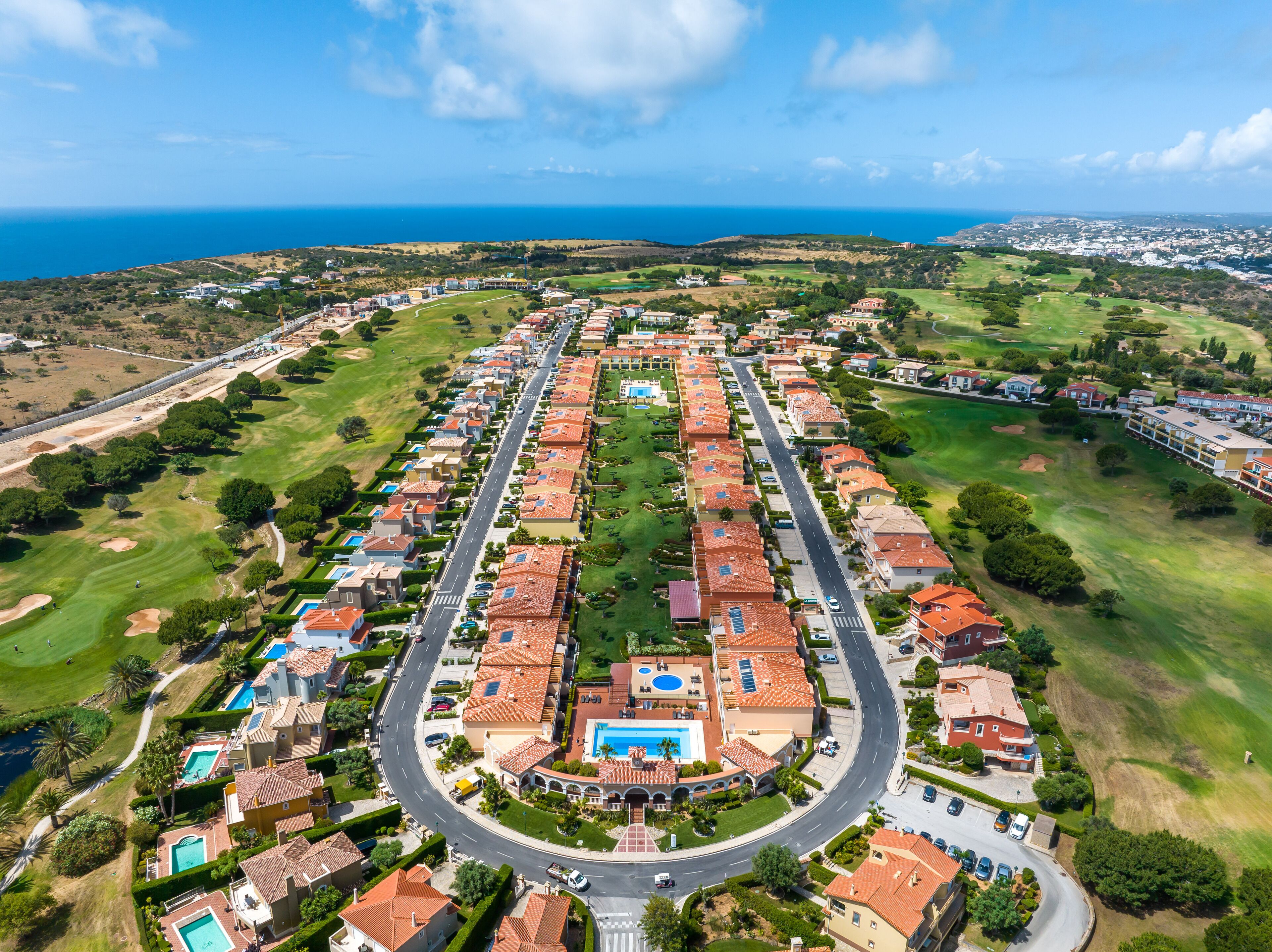 Vista aérea de un barrio residencial con una avenida central, casas con techos de tejas rojas y una piscina grande.