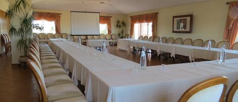A conference room with long tables, chairs, a projector screen, and a large window with curtains.