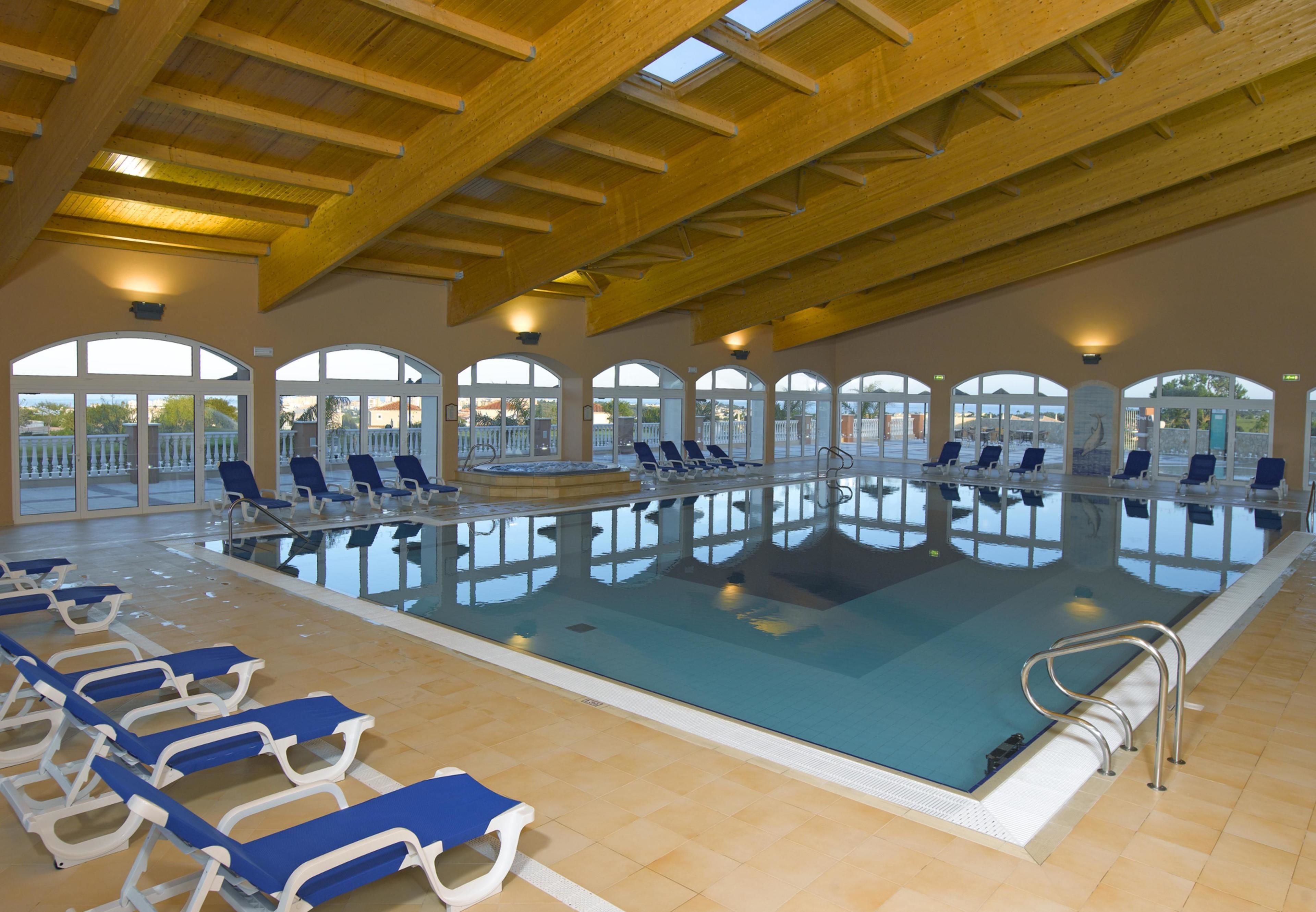 An indoor swimming pool with blue and white lounge chairs, wooden beams, and large arched windows.