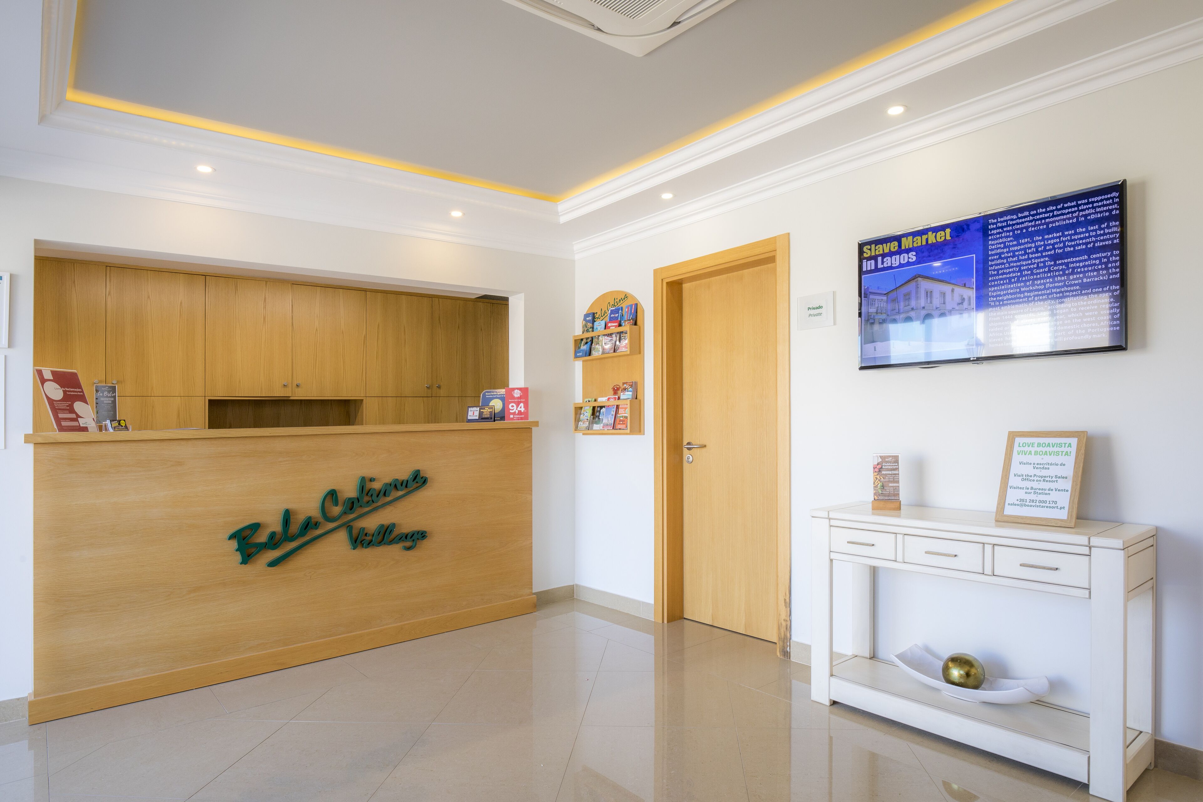 A reception area with a wooden desk labeled "Bela Colonia Vista", a wall-mounted TV displaying "Slave Market in Lagos", and a white console table with a framed certificate.