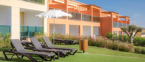 A hotel with outdoor sun loungers, a white umbrella, and a well-maintained lawn.