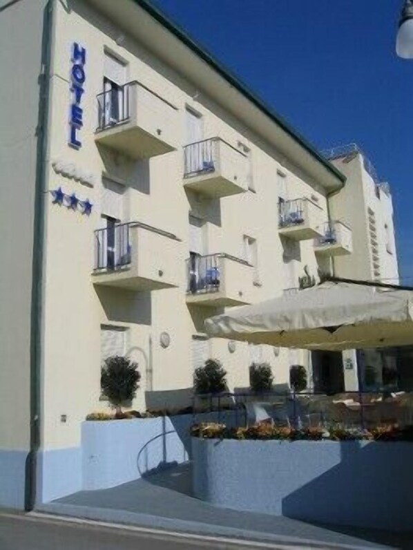 A hotel with a white facade, multiple balconies, and a small outdoor seating area with umbrellas. - Hotel Nina (Bibbona)